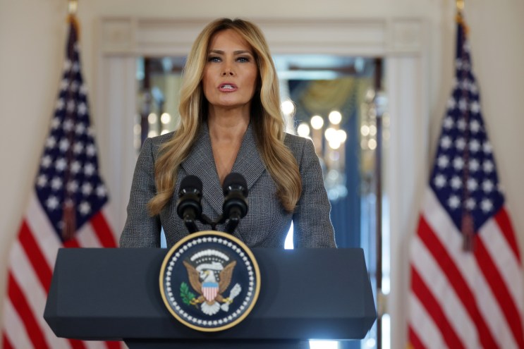 photo of U.S. First Lady Melania Trump at a podium in the White House. Two flags stand on either side of the open door behind her in the background.