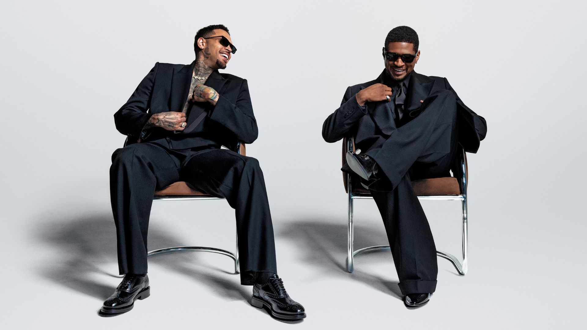 Photo of Usher and Chris Brown sitting on chairs wearing black suits.