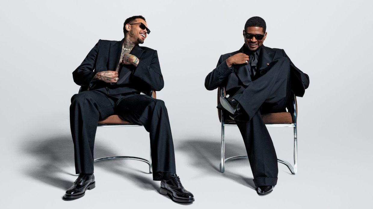 Photo of Usher and Chris Brown sitting on chairs wearing black suits.