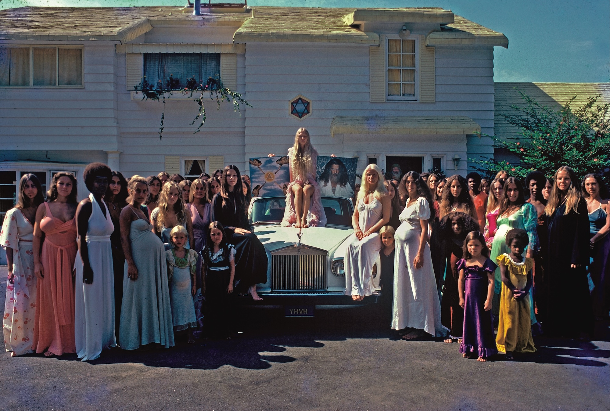 photo depicting a crowd of women around a Rolls-Royce. A woman sits on the car holding a poster depicting a cult leader