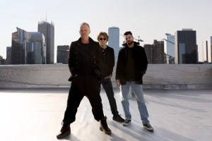 photo of musicians Sting, Dominic Miller, and Chris Maas posing on a rooftop with skyscrapers in the background