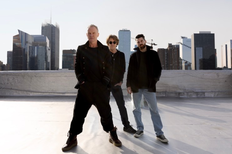 photo of musicians Sting, Dominic Miller, and Chris Maas posing on a rooftop with skyscrapers in the background