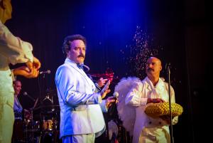 Photo of a an actor onstage paying comedian Alvarez Guedes. Next to him is a man dressed as an angel throwing glitter in the air