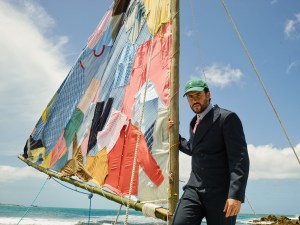 photo of Bad Bunny in a suit and baseball cap standing on a beach next to a sailboat with a sal made of clothes sewn together