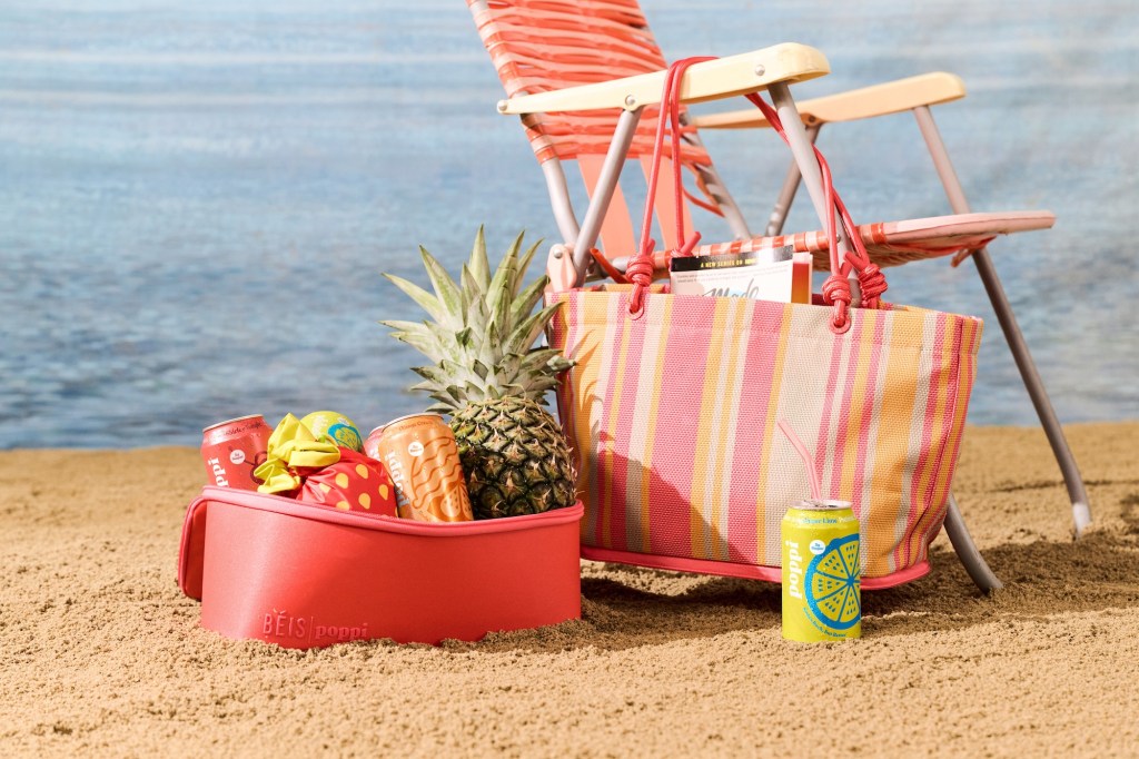photo of a beach chair with a beach bag hanging on it next to a picnic basket with fruits inside in front of the ocean