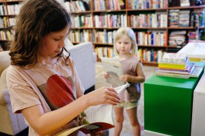 Photo of two girls inside a library. The girl in the foreground reads a book as the younger girl in the background looks at her while holding more books