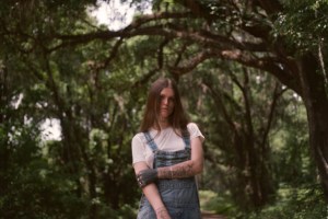 photo of singer Ethel Cain posing in a white t-shirt and denim overalls in front of a path lined with oak trees