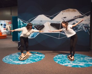 photo of two young girls riding stationary surfboards inside a museum exhibition