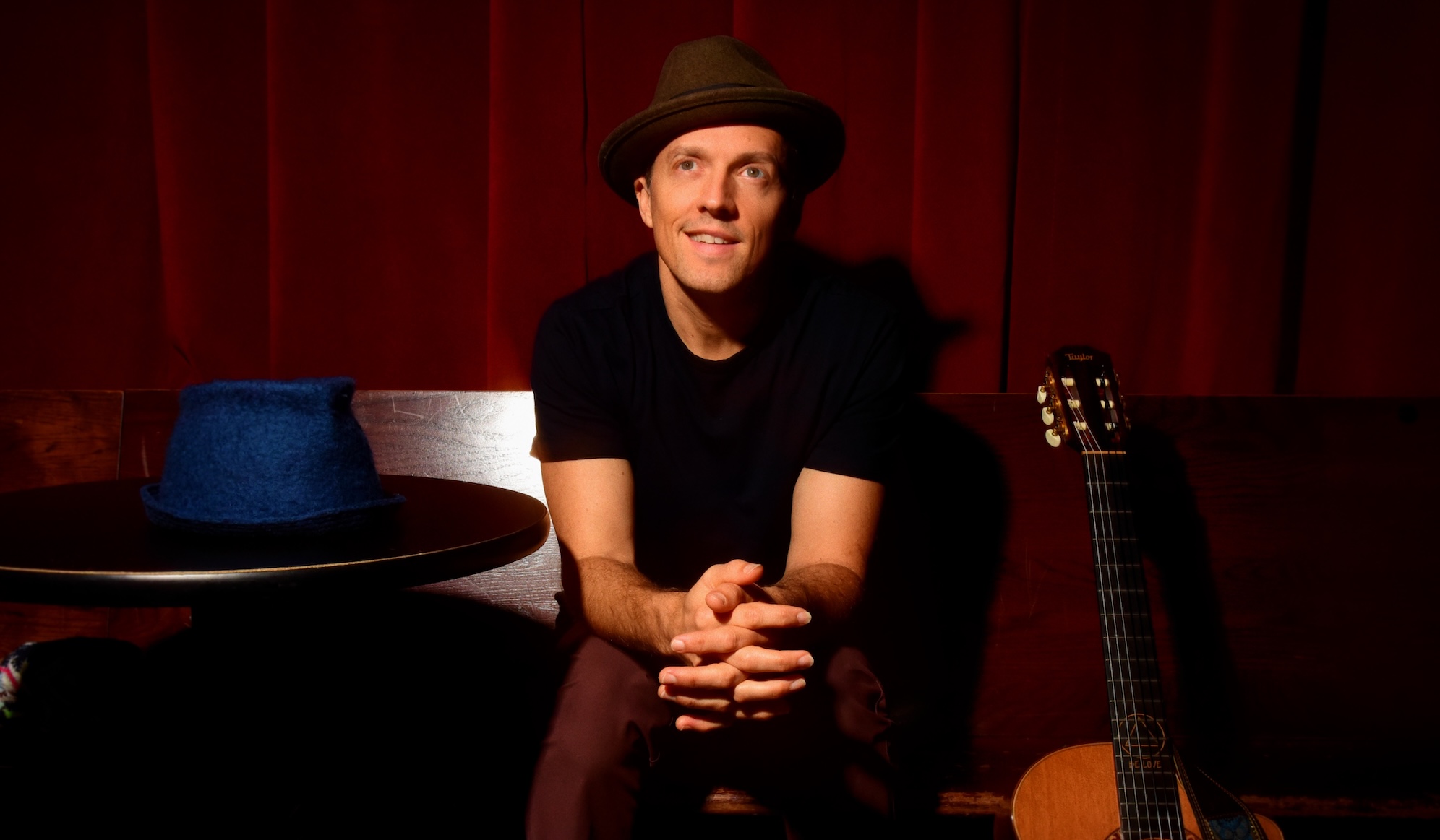 photo of singer/songwriter Jason Mraz in a fedora sitting next to a table with a hat on it to his right and an acoustic guitar to his right