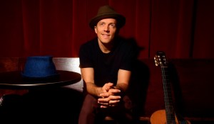 photo of singer/songwriter Jason Mraz in a fedora sitting next to a table with a hat on it to his right and an acoustic guitar to his right