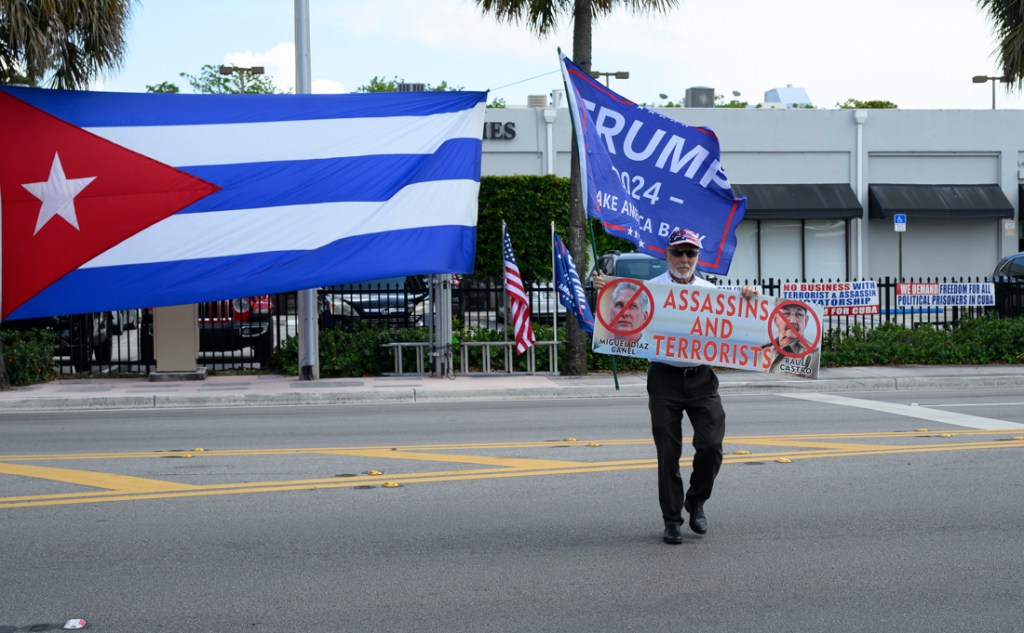 photo of a protester in Miami holding a poster of Cuban leaders Miguel Diaz-Canel and Raul Castro with text reading "assassins and terrorists"