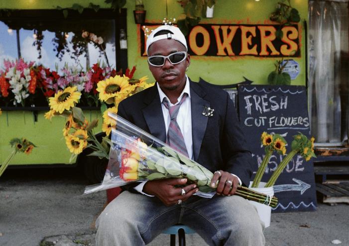 photo of singer Pxtn in a blazer, tie, and denim jeans holding flowers in front of a flower shop