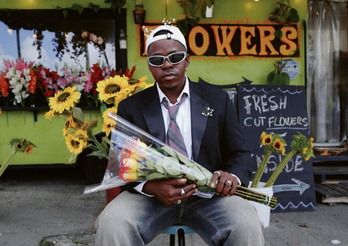 photo of singer Pxtn in a blazer, tie, and denim jeans holding flowers in front of a flower shop