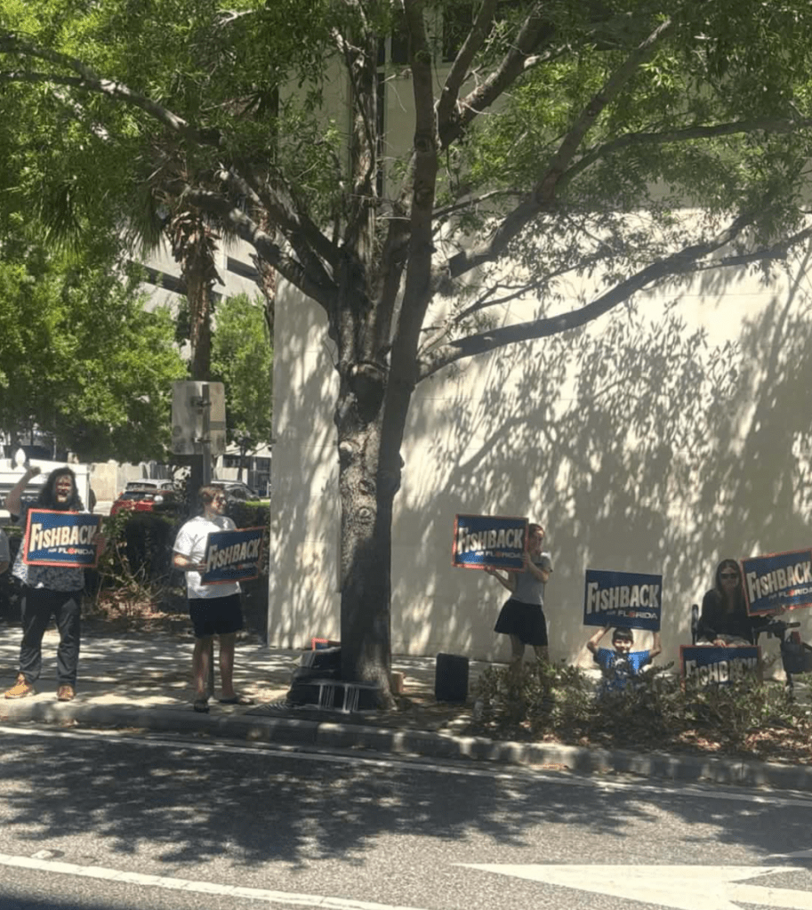 phone photo of four adults and one young boy holding campaign signs reading, "Fishback. outside a building