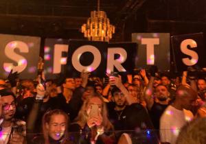 Photo of a dancefloor in Miami with people holding a sign that reads Sports