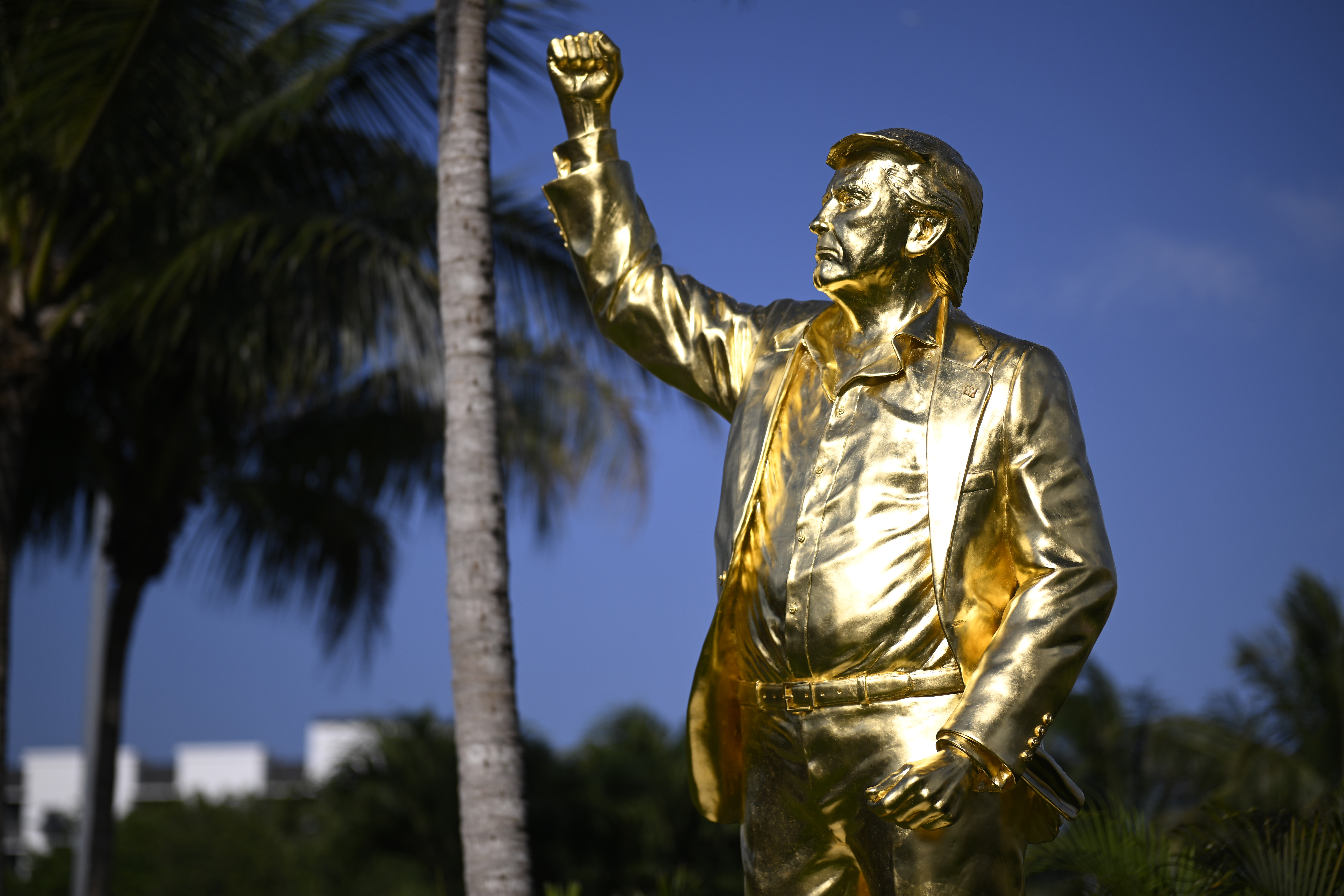 photo of a golden statue of Donald Trump with his fist in the air in front of an evening sky and palm trees