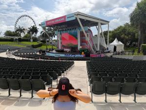 Photo of an outdoor amphitheater with empty seats and a ferris wheel in the background. A person covers their face while showing a baseball cap logo in front of the scene