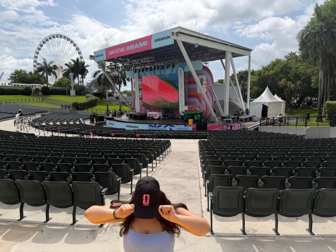 Photo of an outdoor amphitheater with empty seats and a ferris wheel in the background. A person covers their face while showing a baseball cap logo in front of the scene