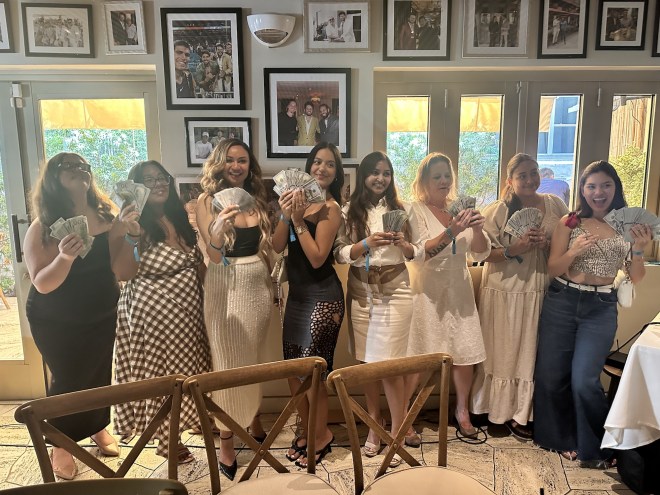 photo of eight women posing together in a row holding up dozens of $100-dollar bills