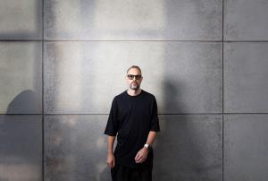 photo of a DJ in shades and a black T-shirt standing in front of a wall of large gray tiles