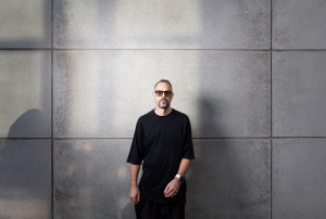 photo of a DJ in shades and a black T-shirt standing in front of a wall of large gray tiles