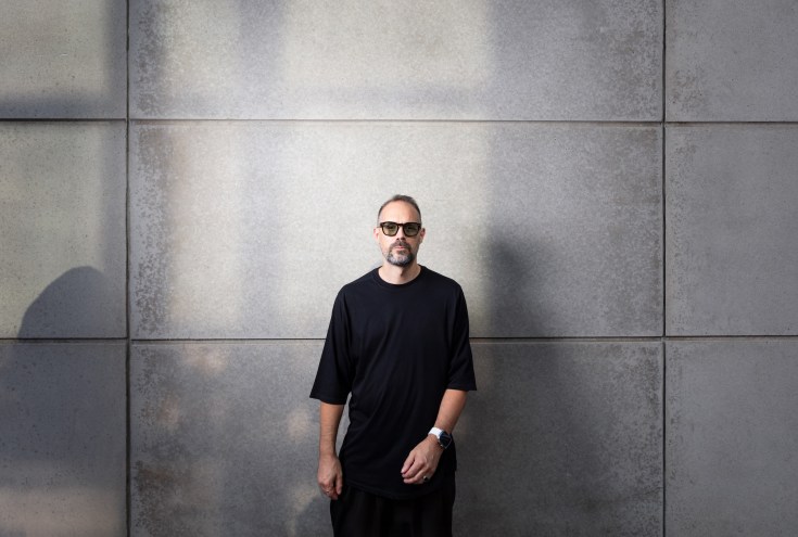 photo of a DJ in shades and a black T-shirt standing in front of a wall of large gray tiles