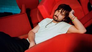 photo of musician Kevin Parker of Tame Impala lounging on a red couch as two other red couches are seen behind him
