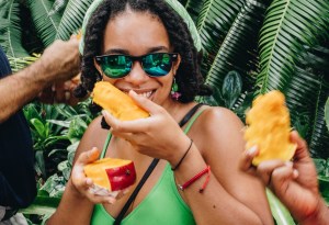 Photo of a woman smiling as she easts a mango, and holds another mango in her other hand. Another person's hand holds a mango next to her