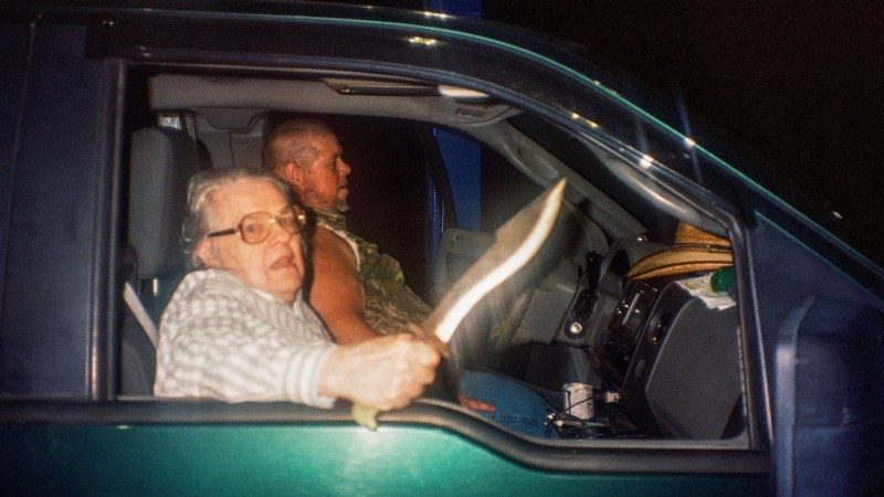 Photo two people inside a car. A man drives and an old woman sits in the passenger seat wielding a knife