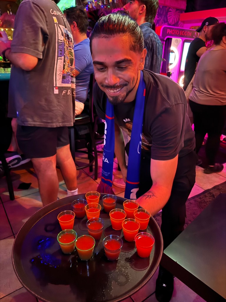a man holds a tray with pink alcoholic shots at a bar