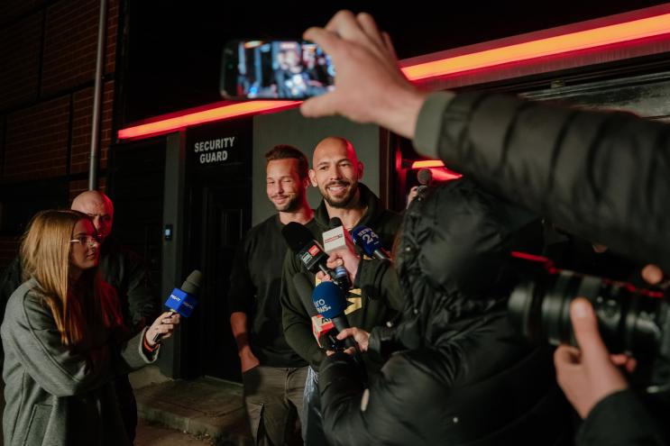 Two men, surrounded by media and onlookers recording on their phones, smile hesitantly for the cameras