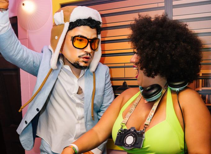Photo of two people behind a DJ booth. The person on the left wears a hyper-realistic Bad Bunny mask and costume. The woman on the right wears a cardboard camera around her neck and turns to the other person laughing