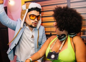 Photo of two people behind a DJ booth. The person on the left wears a hyper-realistic Bad Bunny mask and costume. The woman on the right wears a cardboard camera around her neck and turns to the other person laughing