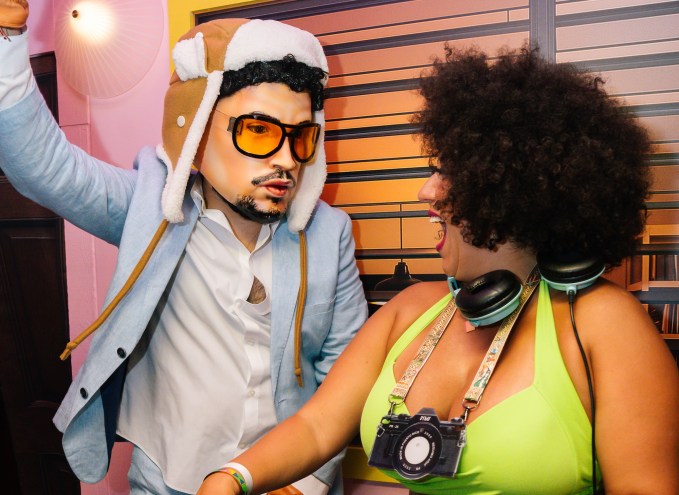 Photo of two people behind a DJ booth. The person on the left wears a hyper-realistic Bad Bunny mask and costume. The woman on the right wears a cardboard camera around her neck and turns to the other person laughing