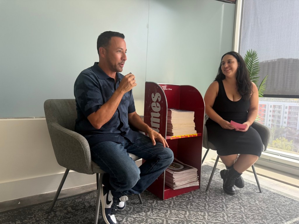 Photo of singer Howie D of the Backstreet Boys recording a podcast episode with host Celia Almeida at the Miami New Times office. A red newspaper rack stacked with papers sits between the two