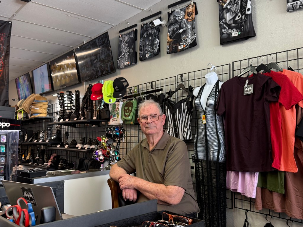 photo of an older man behind the counter of a store selling merchandise geared toward the alternative scene
