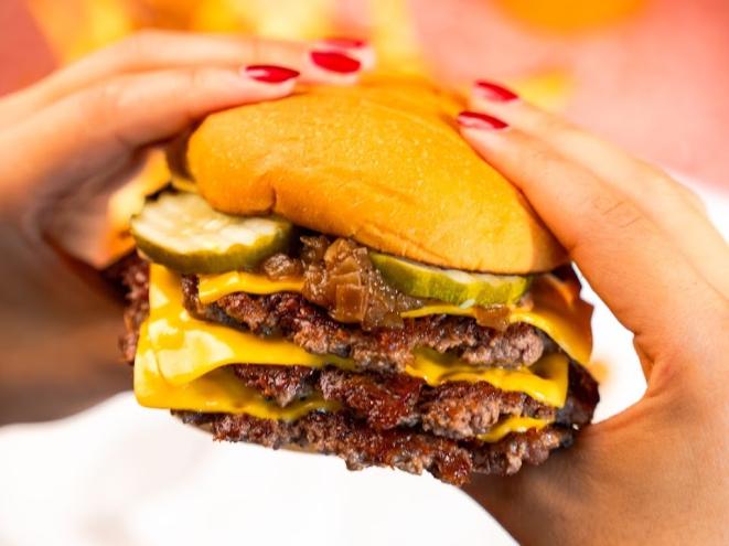 a double cheeseburger with pickles being held by hands with red-painted nails