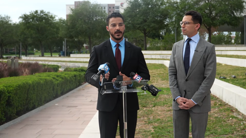A photo of a bearded man in a suit speaking outdoors before a lectern next to a bespectacled man in a suit.