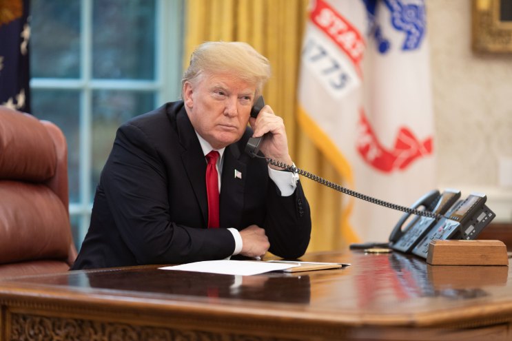 a suited man sits in an office holding a phone to his ear