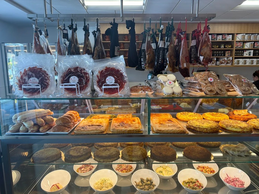 a glass display at a deli featuring baked goods and Spanish ham hanging from the ceiling