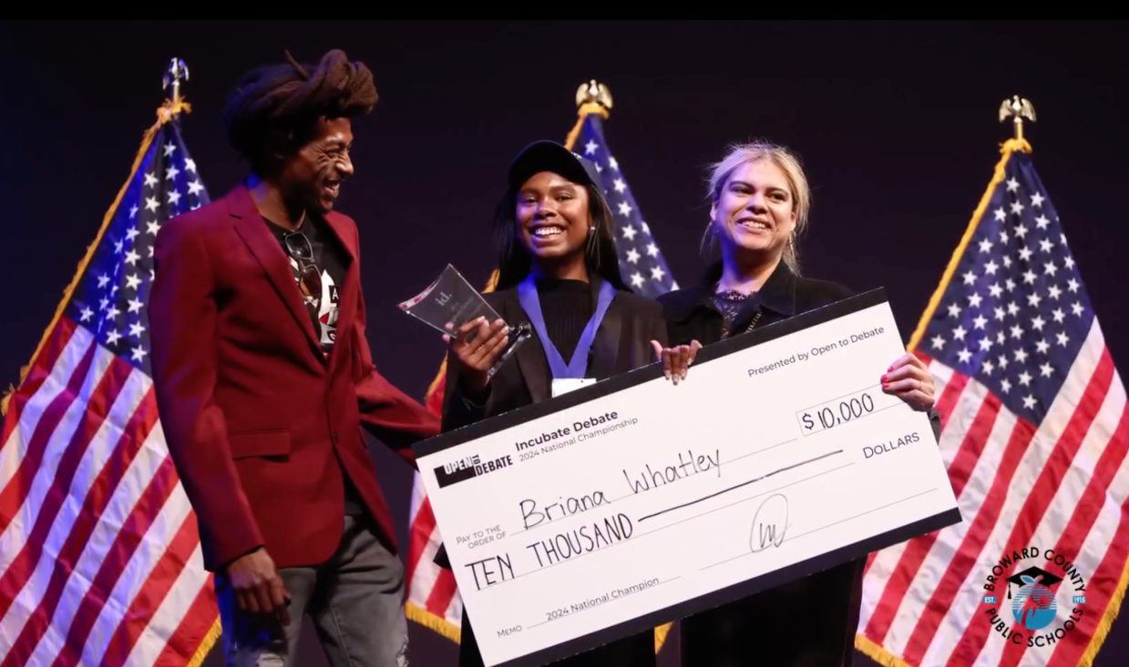 a man in a red blazer and a woman dressed in black had a large check to a high school student debater