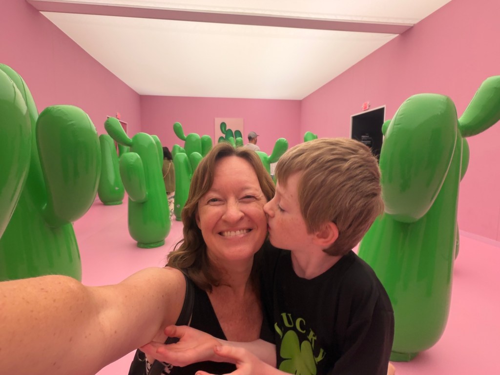 photo of writer Ciara LaVelle taking a selfie with her son giing her a kiss on the cheek inside an immersive museum with cactus sculptures in the background