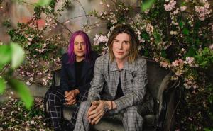 photo of Goo Goo Dolls members John Rzeznik and Robby Takac posing on a couch in slacks and blazers surrounded by flowers and vine