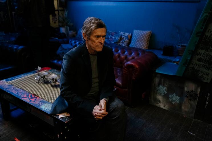 still from the film "Late Fame" depicting Willem Dafoe sitting on a coffee table in the middle of a room filled with furniture and decor