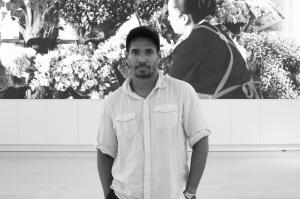 black-and-white photo of artist Luis Manuel Otero Alcántara posing in front of a photograph inside a gallery