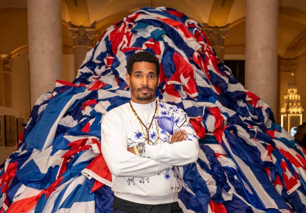 photo of artist Luis Manuel Otero posing in front of a sculpture made of stacked-up Cuban flags