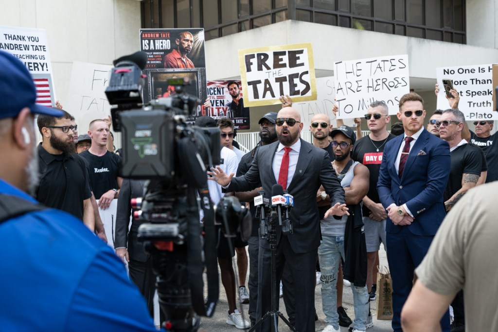 a man in a suit speaks in front of cameras while other men stand around him