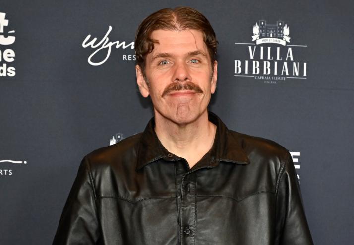 a blue-eyed man with a mustache smiles for a photo on a red carpet