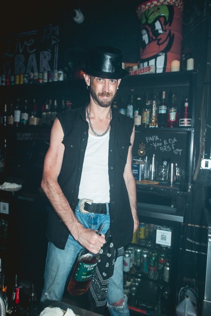 Photo of a man behind the bar holding a bottle of bourbon and wearing a leather hat