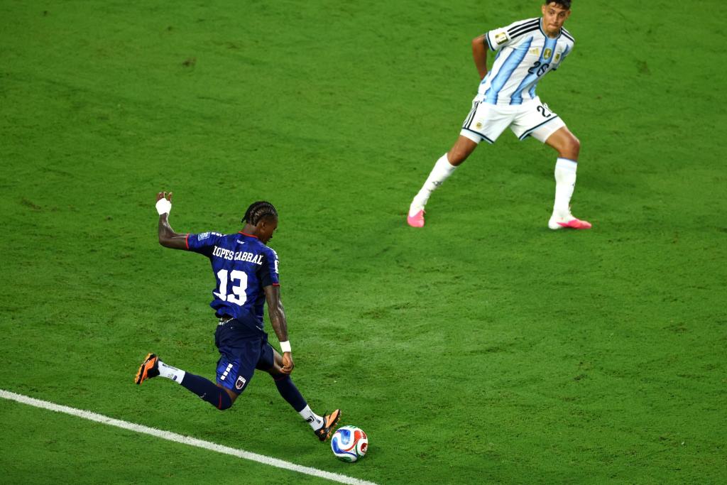 a soccer player in a navy jersey winds up to take a shot as a defender braces to block